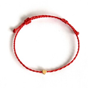 May include: Red string bracelet with a small gold heart charm. The bracelet is adjustable with sliding knots. The heart charm is centered on the red string.