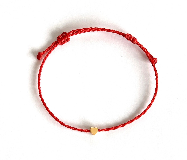 May include: Red string bracelet with a small gold heart charm. The bracelet is adjustable with sliding knots. The heart charm is centered on the red string.