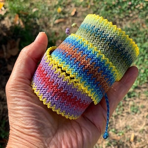 May include: A set of two wide macrame bracelets featuring intricate knotted patterns. The top bracelet displays a gradient of green and blue, while the bottom bracelet showcases a vibrant blend of purple, red, and orange, both edged in bright yellow.