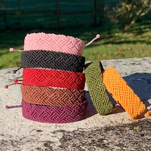 May include: A stack of six macrame bracelets in various colors: pink, black, red, brown, and purple. Two additional bracelets, one olive green and one orange, lean against the stack. The bracelets have a woven chevron pattern and adjustable closures.