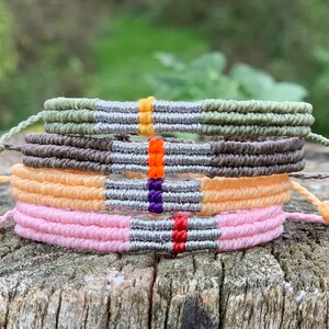 Può includere: Una pila di cinque braccialetti dell'amicizia fatti a mano, su una superficie di legno. I braccialetti sono in vari colori: verde oliva, marrone, pesca e rosa. Ogni braccialetto presenta un accento argentato e una striscia colorata.