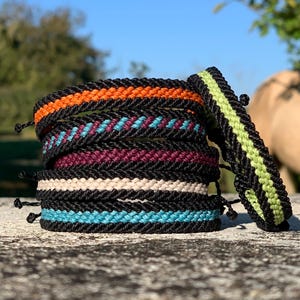 Puede incluir: Una pila de pulseras tejidas en varios colores, incluyendo naranja, turquesa, burdeos y crema, con detalles en negro. Una pulsera tiene una franja verde lima. Las pulseras se muestran sobre una superficie texturizada.