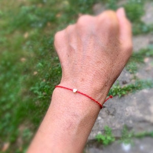 May include: Red string bracelet with a small gold heart charm. The bracelet is tied around a wrist, with the background blurred, suggesting an outdoor setting. The heart charm is centered on the red string.