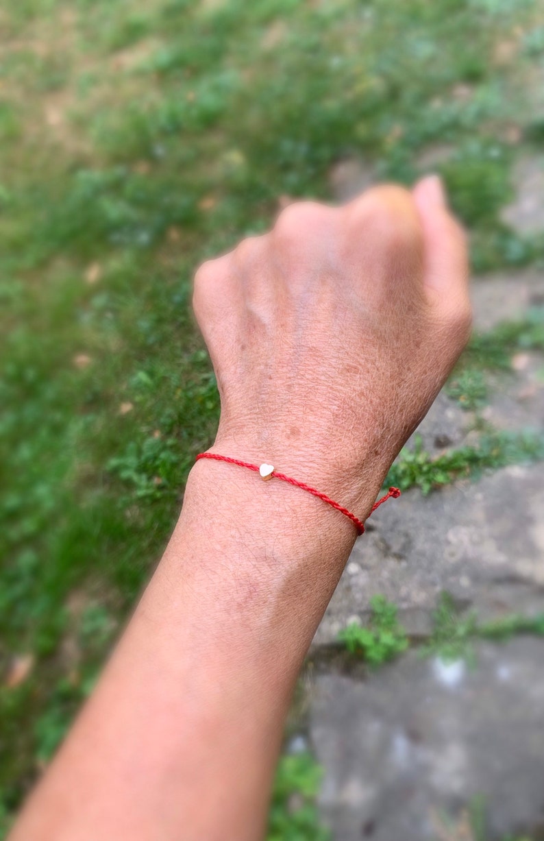 May include: Red string bracelet with a small gold heart charm. The bracelet is tied around a wrist, with the background blurred, suggesting an outdoor setting. The heart charm is centered on the red string.