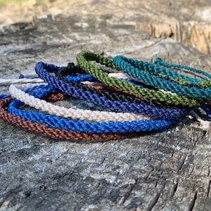 May include: A selection of braided rope bracelets in shades of brown, blue, green, and white. The bracelets are displayed on a weathered wooden surface, highlighting their handcrafted nature and earthy tones. These bracelets are suitable for daily use.