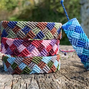 Peut inclure: Quatre bracelets en macramé de couleurs variées, empilés sur une surface en bois. Les bracelets présentent un motif tissé dans des tons de bleu, vert, marron, rose et violet. Un seul bracelet bleu est debout, mettant en valeur le design.