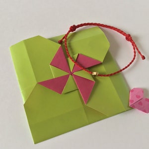May include: A lime green origami gift box with a pink and red origami flower and a red string bracelet with a gold bead. A small pink heart-shaped origami is also present.