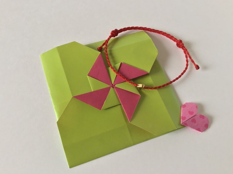 May include: A lime green origami gift box with a pink and red origami flower and a red string bracelet with a gold bead. A small pink heart-shaped origami is also present.