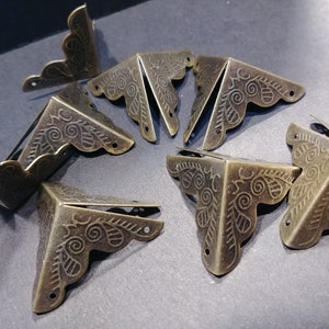 May include: A collection of antique bronze-colored metal corner protectors. Each L-shaped piece features an ornate, scrolled design along the outer edge and small holes for attachment. These decorative hardware pieces are ideal for crafting and scrapbooking projects.