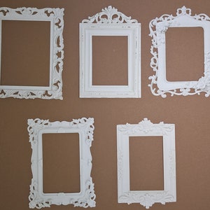 May include: Five white, ornate picture frames with detailed designs are displayed against a brown backdrop. The frames have different shapes and decorative elements, suggesting a vintage style. Each frame is empty, ready for a photograph or artwork.