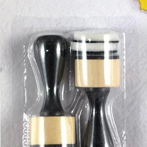 May include: Two ink applicators in a clear plastic package. Each applicator has a black handle and a round, light-colored foam head with black and white stripes. The packaging includes the text "Ink Applicator round 2pcs" and a logo.