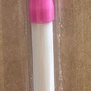 May include: A double-ended dotting tool for nail art, featuring a white and pink handle. The tool has a metal tip on each end, one with a small ball and the other with a larger ball. It is packaged in clear plastic.