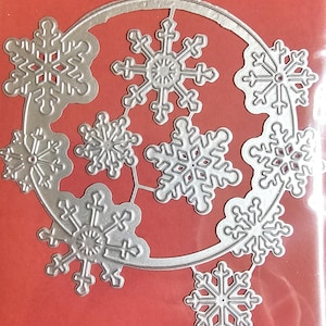 May include: A silver metal snowflake die cut with eight detailed snowflakes arranged in a circular pattern. The die cut is on a red background, perfect for holiday crafts and decorations.