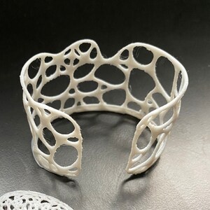 Pode incluir: Um conjunto de três peças de joalheria brancas impressas em 3D. O conjunto inclui uma pulseira e dois brincos. A pulseira e os brincos são feitos de um design delicado e vazado com um padrão circular.