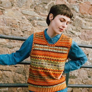 Könnte beinhalten: Eine farbenfrohe, gemusterte Strickweste in Orange-, Gelb- und Grüntönen. Die Weste wird über einem blauen Jeanshemd und einer beigen Hose getragen. Der Hintergrund zeigt eine Steinmauer und einen Metallzaun.