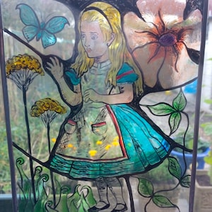 May include: A stained glass window featuring Alice from Alice in Wonderland. Alice is wearing a blue and white dress with a red sash. She is standing in a garden with flowers and a butterfly.