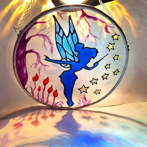 May include: A stained glass window hanging with a blue fairy with wings, holding a wand, surrounded by yellow stars and a purple tree with red flowers.