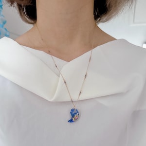 Puede incluir: Un collar de cadena dorada con un colgante azul y blanco con forma de media luna. El colgante tiene un diseño detallado con pequeños detalles coloridos.