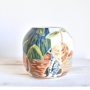 May include: A ceramic vase with a hand-painted design featuring seashells, plants, and a coastal scene. The vase has a rounded shape and is primarily white, with accents of blue, green, brown, and orange. The vase is sitting on a white surface.