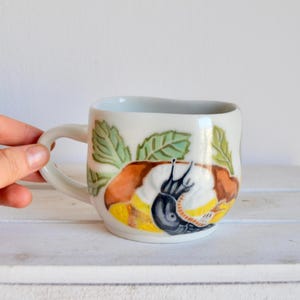 Könnte beinhalten: Weiße Keramiktasse mit handgemaltem Design einer Schnecke auf einem Blatt. Die Schnecke ist schwarz mit einer braunen Schale und von grünen Blättern umgeben.