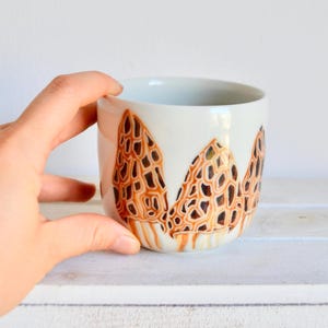 Può includere: Tazza in ceramica bianca con un design a fungo marrone e arancione. La tazza è dipinta a mano con un motivo dettagliato.
