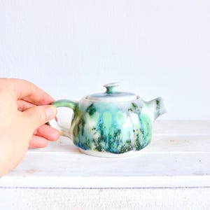 Op de afbeelding: Kleine keramische theepot met deksel, handvat en tuit. De theepot is beschilderd met een mix van turquoise, groene en blauwe tinten, met gouden spikkels. Een hand houdt de theepot vast.