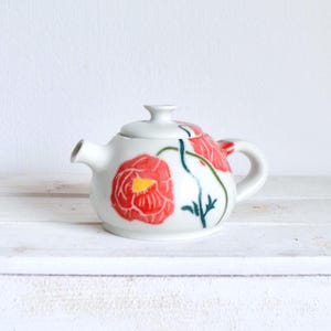 Könnte beinhalten: Kleine, weiße Keramik-Teekanne mit Deckel, verziert mit roten Mohnblumen und grünen Stielen. Die Teekanne hat einen kleinen Ausguss und einen Henkel. Die Mohnblumen haben gelbe Zentren. Die Teekanne steht auf einer weißen Oberfläche.