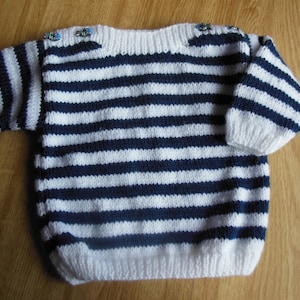 Peut inclure: Un pull en tricot rayé blanc et bleu marine avec un col bateau et des boutons décoratifs sur les épaules.