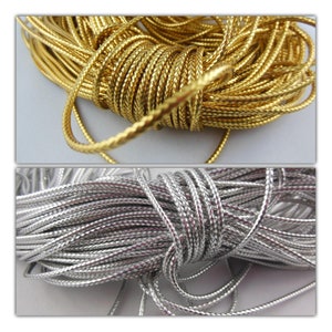 Può includere: Primo piano di due matasse di cordoncino intrecciato. Uno è di colore oro brillante, l'altro argento. I cordoncini sono fittamente intrecciati, creando una superficie strutturata. I cordoncini sono probabilmente utilizzati per la creazione di gioielli o lavori artigianali.