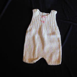 Peut inclure: Un body bébé blanc en tricot avec des nœuds roses. Le body a un motif moucheté et est sans manches.