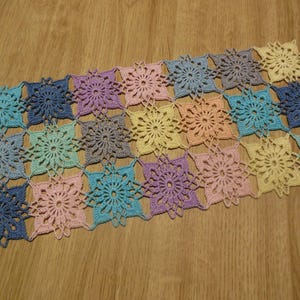 May include: A colorful, handmade crochet table runner. The runner is made of individual floral squares in shades of blue, green, pink, purple, and yellow. The crochet work is detailed and intricate, creating a decorative textile.
