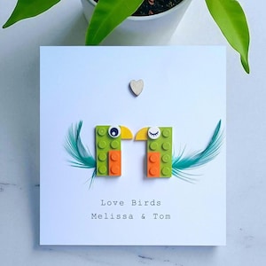 Puede incluir: Tarjeta de felicitación blanca con dos pájaros hechos con bloques verdes y naranjas, con picos amarillos y ojos azules. La tarjeta incluye el texto "Love Birds" y los nombres "Melissa & Tom", con un pequeño corazón de madera y plumas color teal.