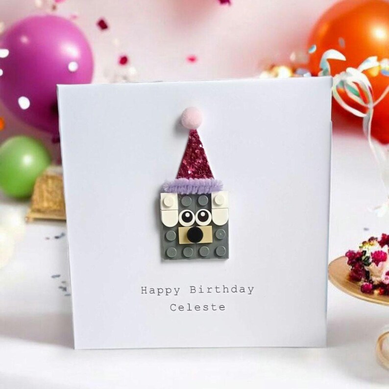 May include: A white card with a gray Lego figure wearing a pink and purple party hat. The card says "Happy Birthday Celeste".