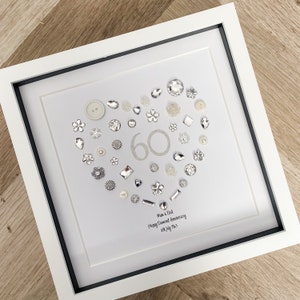 May include: A white frame with a white background and a silver heart made of buttons, rhinestones, and other embellishments. The number 60 is in the centre of the heart, and the text "Mom & Dad Happy Diamond Anniversary 18th July 2023" is below the heart.