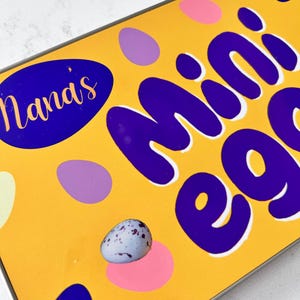 May include: A yellow tin with the words "Nana's Mini Eggs" in purple lettering. The tin is decorated with purple, pink, and white polka dots.