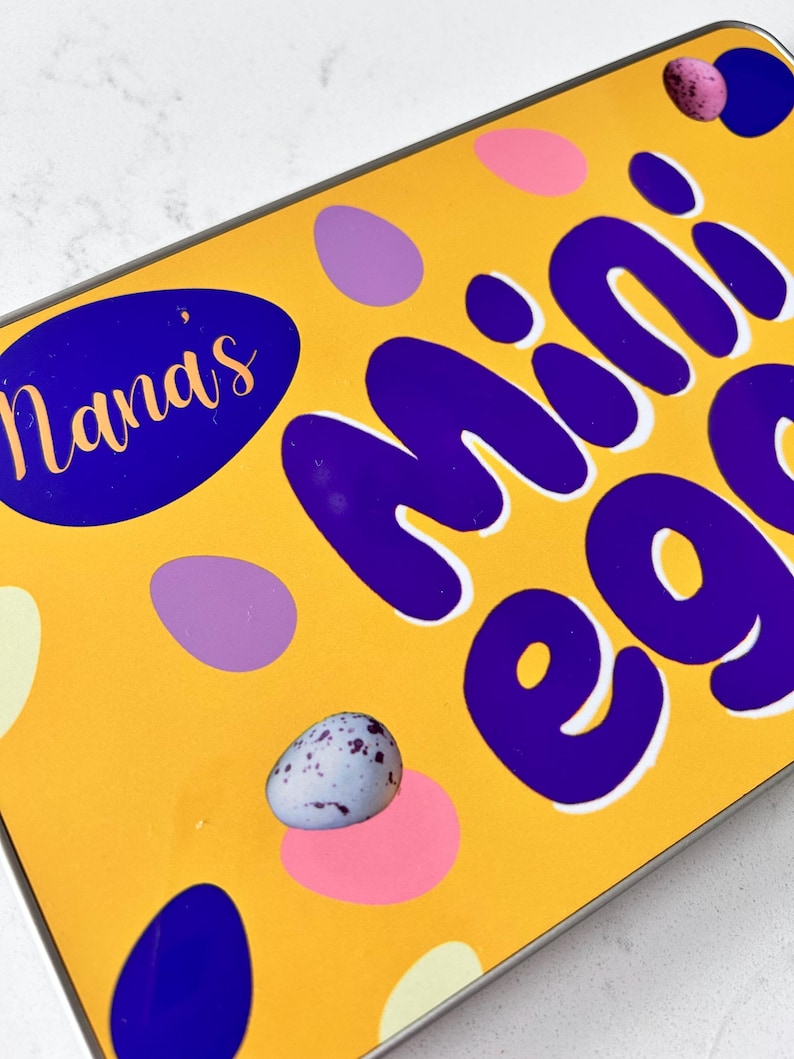 May include: A yellow tin with the words "Nana's Mini Eggs" in purple lettering. The tin is decorated with purple, pink, and white polka dots.