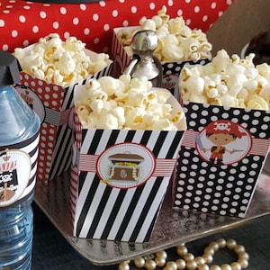 May include: Four black and white striped popcorn boxes with red accents and pirate themed designs. The boxes are filled with popcorn and are sitting on a silver tray.