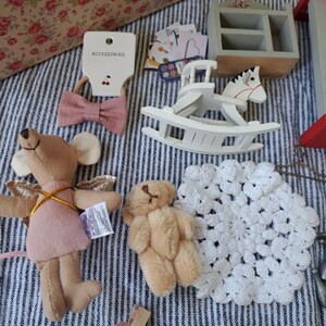Op de afbeelding: Een verzameling miniatuurspeelgoed en accessoires, waaronder een roze strik haarband, een wit schommelpaard, een stoffen muis met vleugels, een kleine teddybeer en een wit gehaakt kleedje. De items zijn gerangschikt op een gestreepte stof.