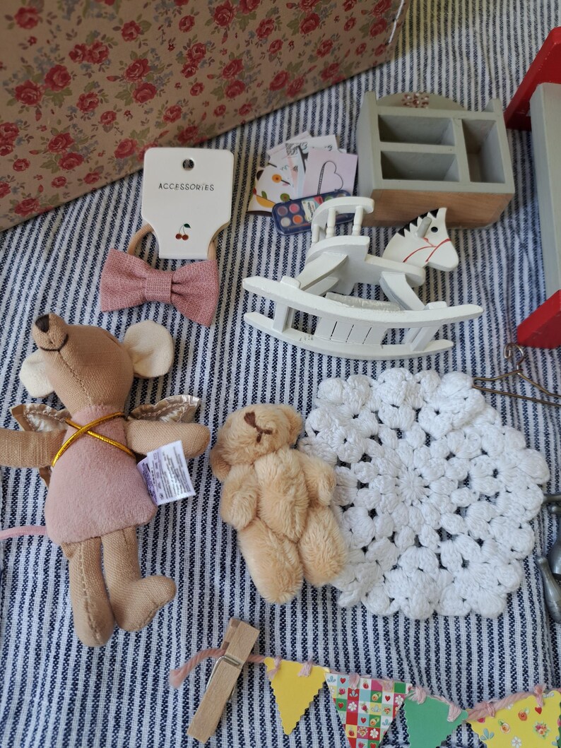 Op de afbeelding: Een verzameling miniatuurspeelgoed en accessoires, waaronder een roze strik haarband, een wit schommelpaard, een stoffen muis met vleugels, een kleine teddybeer en een wit gehaakt kleedje. De items zijn gerangschikt op een gestreepte stof.