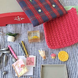 Op de afbeelding: Een verzameling miniatuurartikelen, waaronder een rood-blauwe geruite stof, een roze gebreid vierkant, potloden, een miniatuurboek, een dienblad en kleine voedselreplica's. De items zijn gerangschikt op een blauw-wit gestreepte stof.
