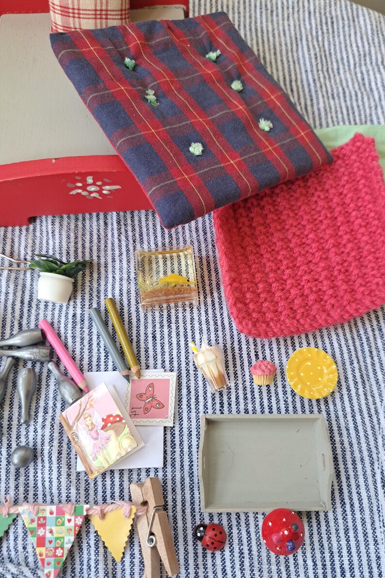 Op de afbeelding: Een verzameling miniatuurartikelen, waaronder een rood-blauwe geruite stof, een roze gebreid vierkant, potloden, een miniatuurboek, een dienblad en kleine voedselreplica's. De items zijn gerangschikt op een blauw-wit gestreepte stof.