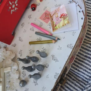 Op de afbeelding: Een miniatuursc&egrave;ne in een decoratieve doos, met een rood geschilderd houten bed, een fee&euml;nillustratie, potloden en kleine grijze kegelvormige objecten. Een lieveheersbeestje en een paddenstoelvormige decoratie dragen bij aan het grillige thema. De doos heeft een bloemmotief interieur.