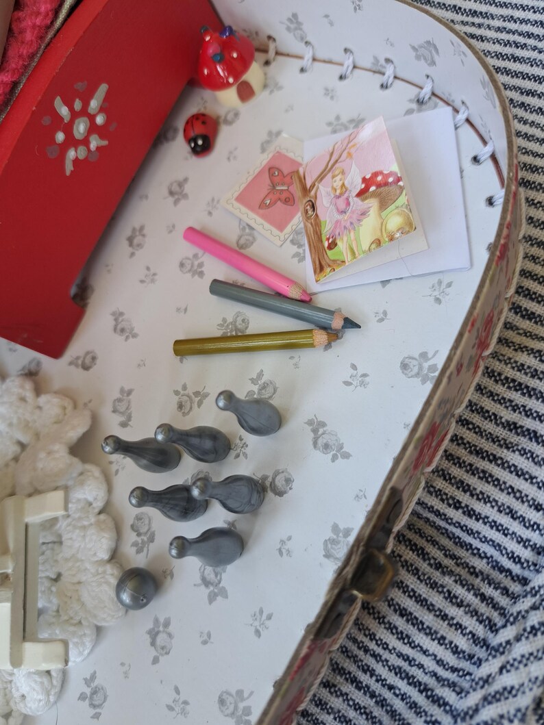 Op de afbeelding: Een miniatuursc&egrave;ne in een decoratieve doos, met een rood geschilderd houten bed, een fee&euml;nillustratie, potloden en kleine grijze kegelvormige objecten. Een lieveheersbeestje en een paddenstoelvormige decoratie dragen bij aan het grillige thema. De doos heeft een bloemmotief interieur.