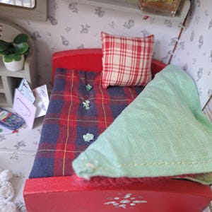 Op de afbeelding: Een miniatuur rood bed met een geruite deken en een rood-wit geblokt kussen. Een lichtgroene deken is over het bed gevouwen. Het bed staat in een poppenhuisslaapkamer met bloemenbehang.