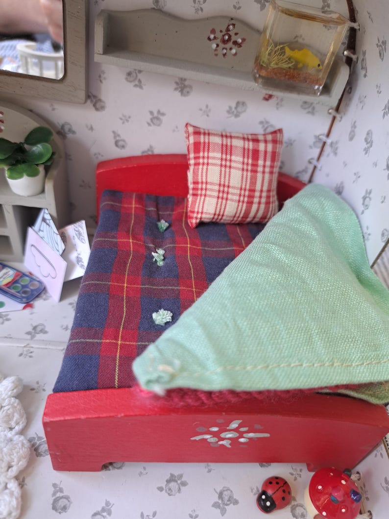 Op de afbeelding: Een miniatuur rood bed met een geruite deken en een rood-wit geblokt kussen. Een lichtgroene deken is over het bed gevouwen. Het bed staat in een poppenhuisslaapkamer met bloemenbehang.