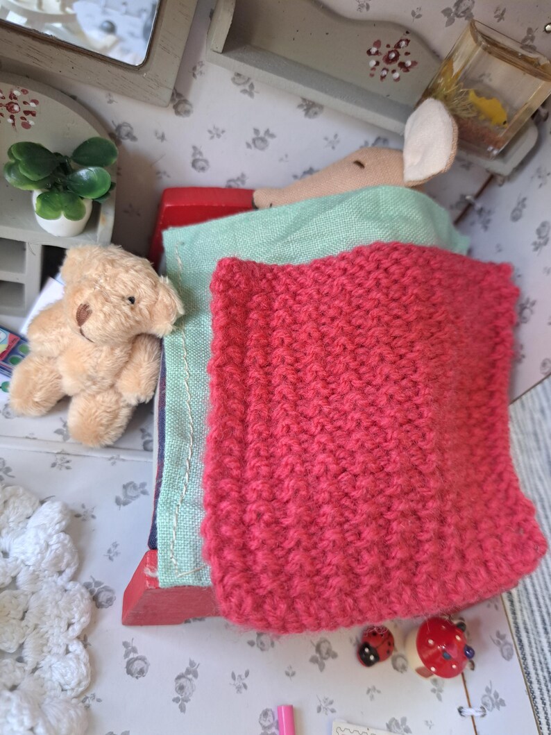 Op de afbeelding: Een miniatuur poppenhuis sc&egrave;ne met een teddybeer, een roze gebreide deken en een groene doek. Een kleine muispop is op de achtergrond. Decoratieve lieveheersbeestjes knopen en een potplant voegen details toe.