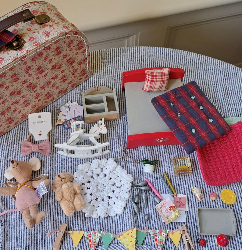 Op de afbeelding: Een verzameling miniatuurspeelgoed en accessoires, waaronder een rood-witte bloemenkoffer, een rood bed met geruite beddengoed, een wit schommelpaard en een pluchen muis. Andere items zijn een doily, een banner en een kleine vogelkooi.
