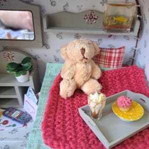 Op de afbeelding: Miniatuur poppenhuis slaapkamer sc&egrave;ne met een bruine teddybeer op een rode gebreide deken. Een klein dienblad bevat een cupcake en een dessertdrankje. Een aquarium en een spiegel zijn ook zichtbaar.