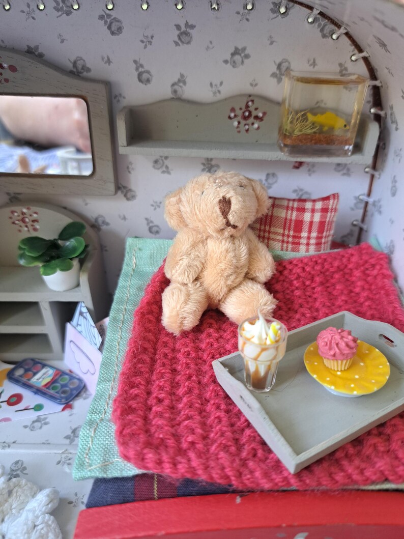 Op de afbeelding: Miniatuur poppenhuis slaapkamer sc&egrave;ne met een bruine teddybeer op een rode gebreide deken. Een klein dienblad bevat een cupcake en een dessertdrankje. Een aquarium en een spiegel zijn ook zichtbaar.