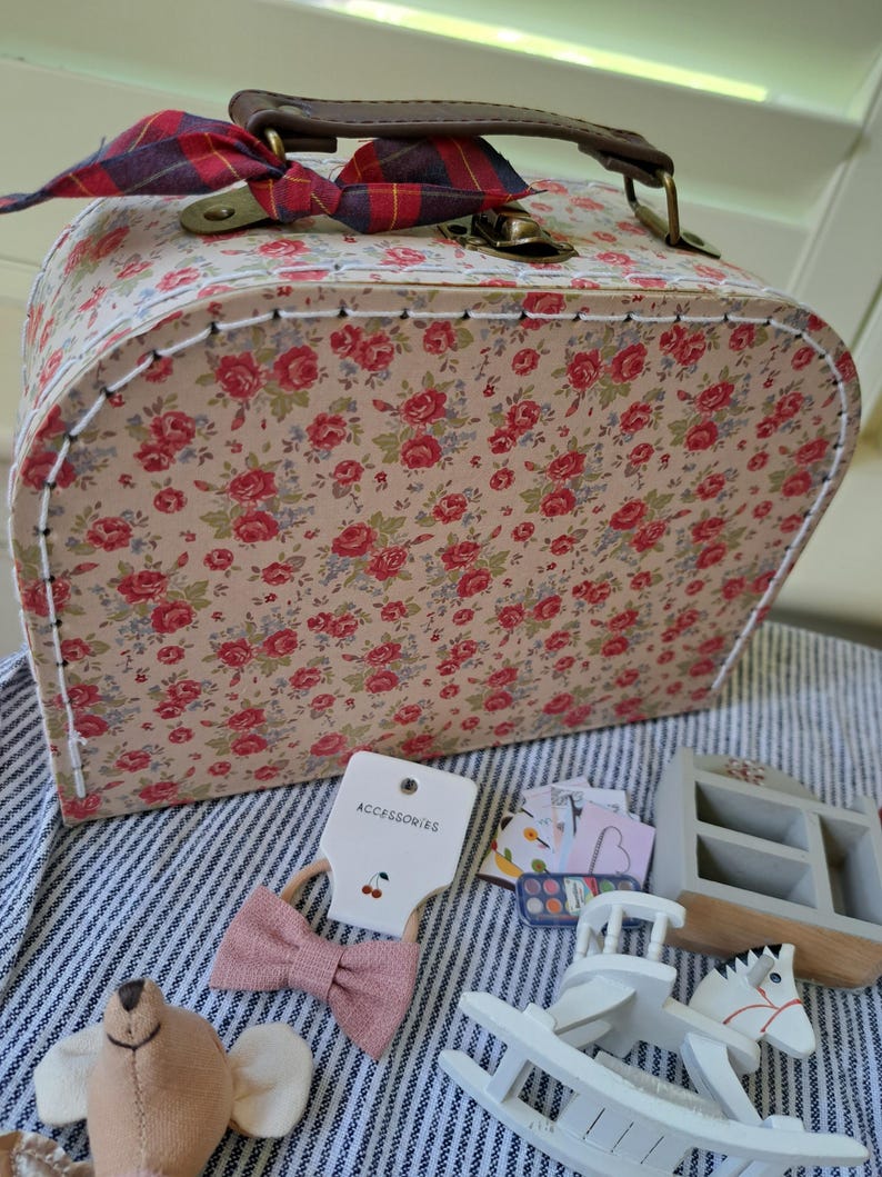 Op de afbeelding: Een bloemenpatroon koffer met een bruin handvat en een geruite strik. Accessoires zijn onder meer een roze strik, een speelgoed schommelpaard en een kleine houten vitrine. De koffer is versierd met rode rozen.
